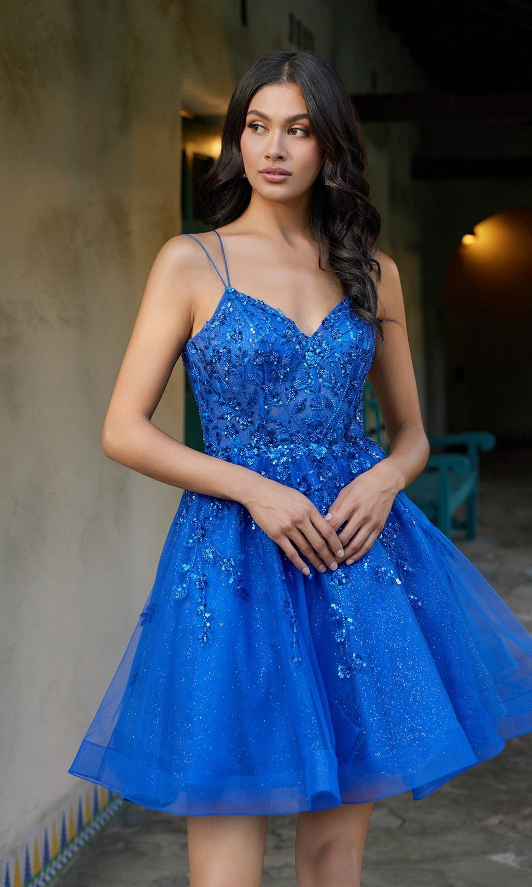 A woman stands indoors with her hands clasped in front, looking to the side, wearing the Juliet JT988N short homecoming dress in royal blue featuring a sequin-embellished corset bodice and sleeveless design.
